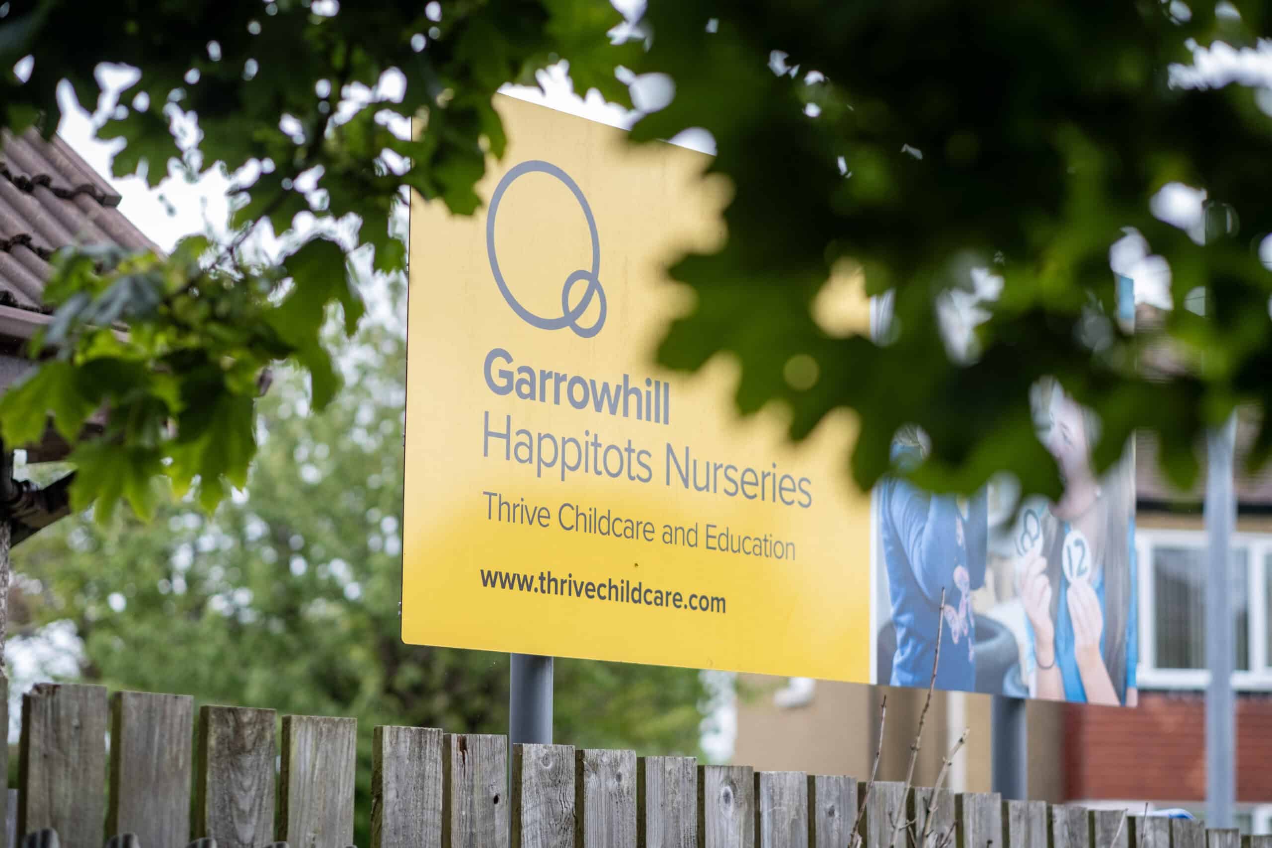 Bright yellow sign for Garrowhill Happitots Nurseries, part of Thrive Childcare and Education, surrounded by green leaves, providing information about childcare services in the local community.