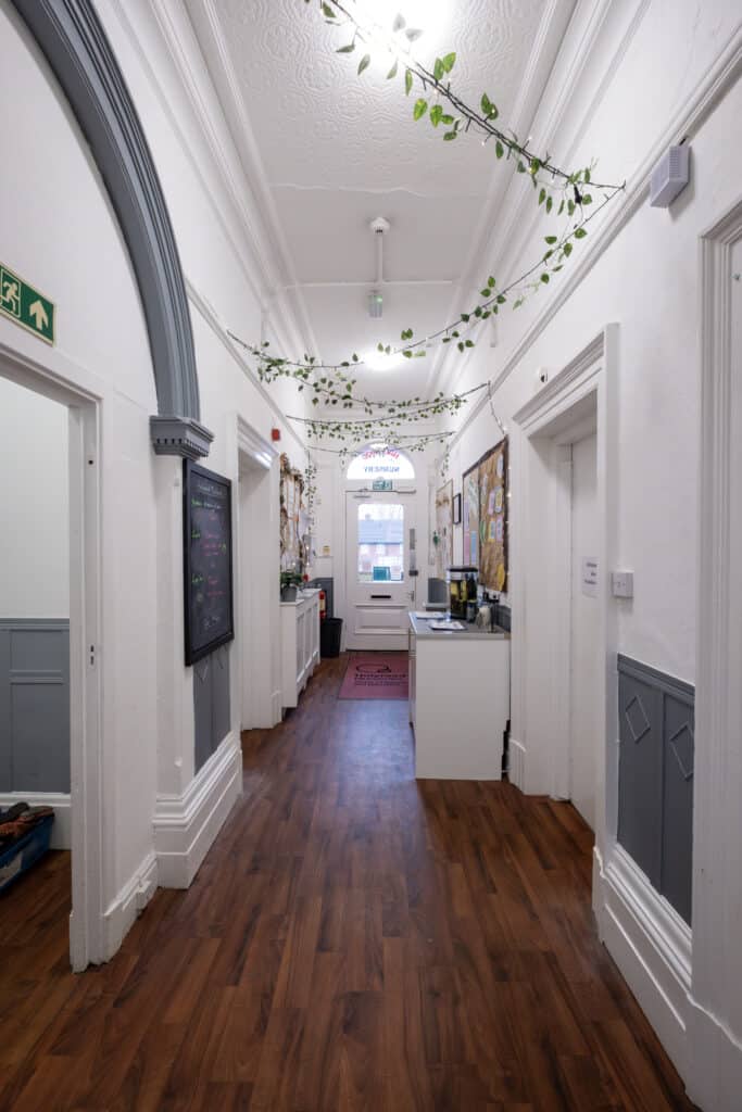 Indoor corridor of Thrive Childcare with white walls, wooden flooring, and decorative foliage on the ceiling, creating a welcoming environment for children and families.