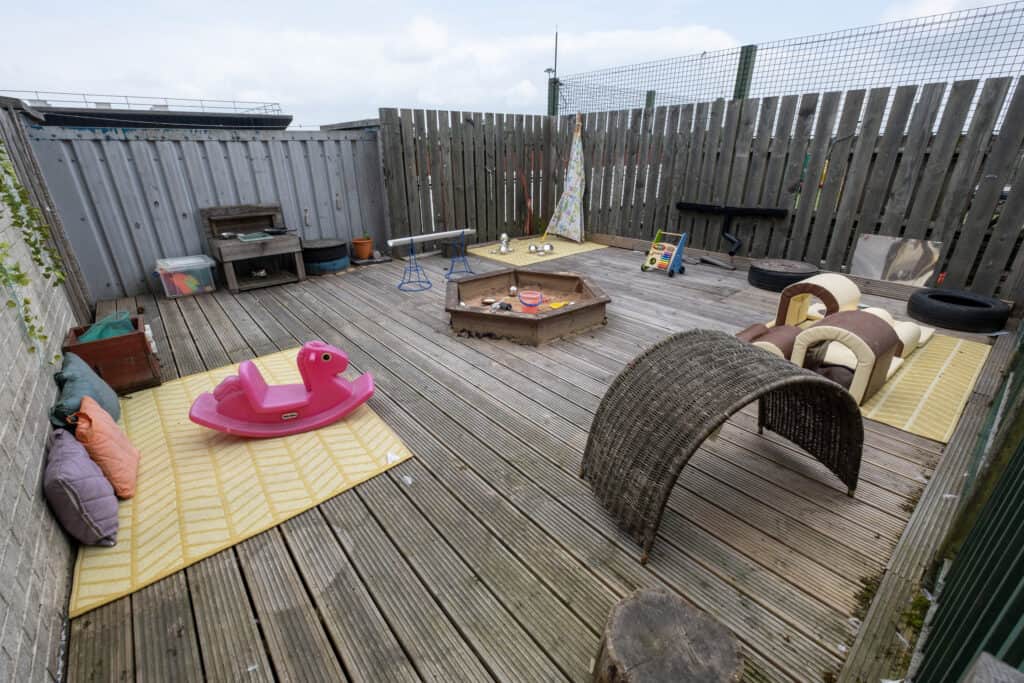 Bright outdoor toddler play area at Thrive Childcare with sandbox, rocking horse, cushions, and toys, providing a safe and engaging environment for young children to explore and play.