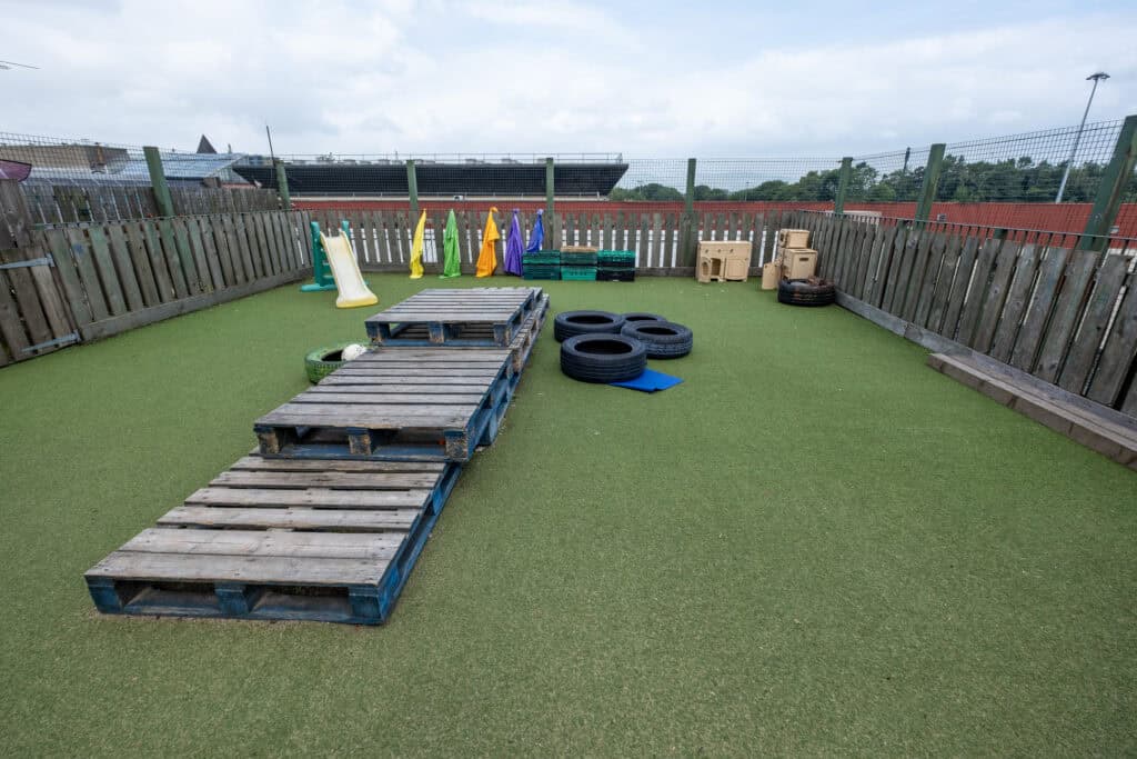 Bright outdoor play area at Thrive Childcare with artificial grass, children's slides, climbing pallets, tyres, and sensory toys, perfect for early childhood play and development.