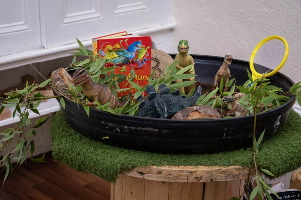 Dinosaur-themed sensory bin with toy dinosaurs, green leaves, a children's book, and a yellow magnifying glass in a childcare setting for imaginative play and learning.