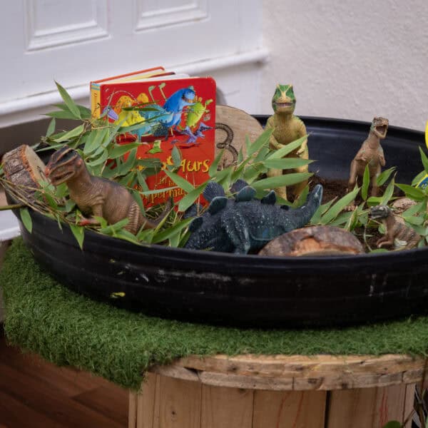 Dinosaur-themed sensory bin with toy dinosaurs, green leaves, a children's book, and a yellow magnifying glass in a childcare setting for imaginative play and learning.
