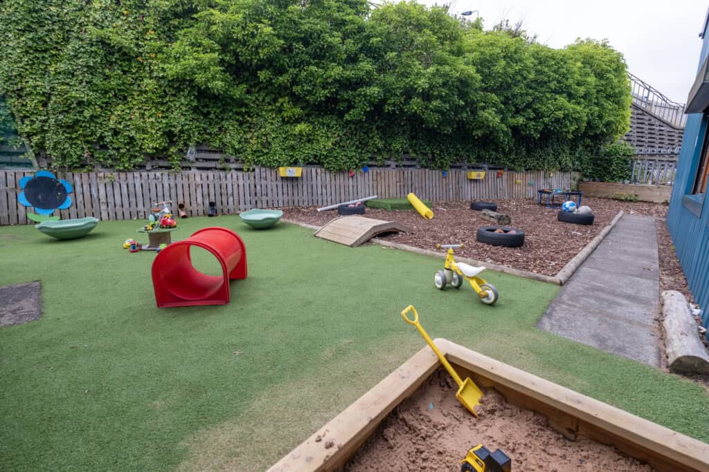 Bright outdoor children's play area at Thrive Childcare with toys, climbing equipment, tyres, sandbox, and green artificial grass, perfect for early childhood play and development.