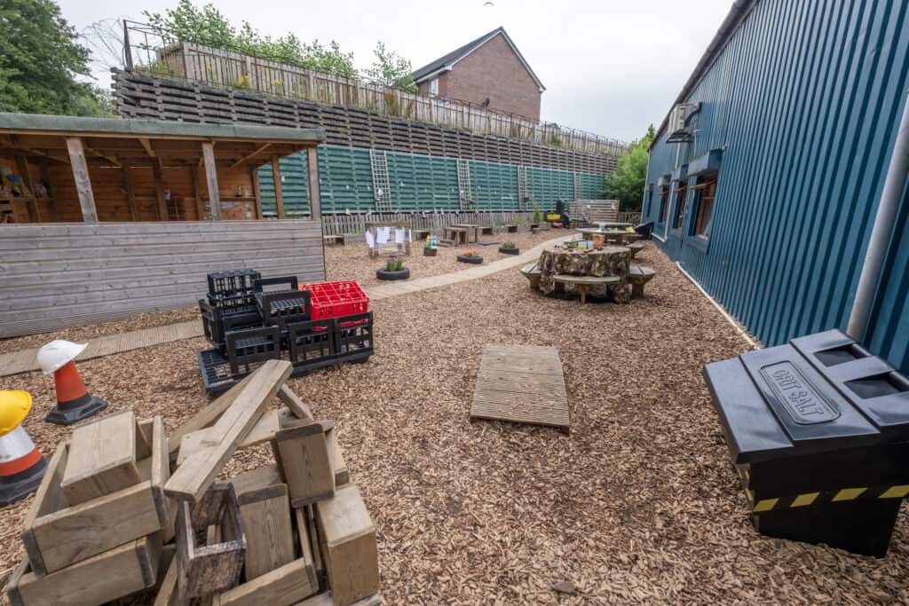 Outdoor play area at Thrive Childcare, featuring natural wooden structures, toy crates, safety cones, and a large blue building, providing a safe and engaging environment for children's activities.