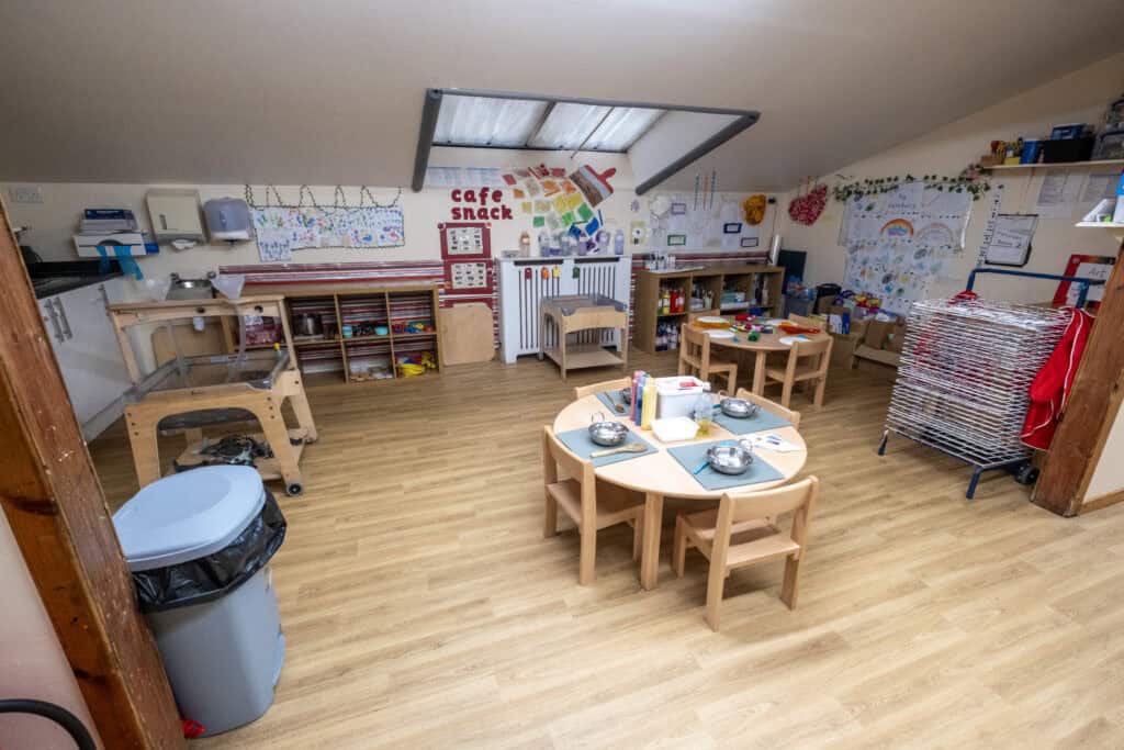 Bright indoor childcare playroom with wooden tables, art supplies, toys, and a cosy learning environment at Thrive Childcare, promoting early childhood development and fun educational activities.