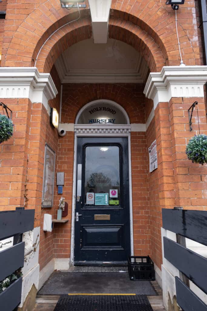 Bright red brick nursery entrance with "Holyrood Nursery" sign above door, decorative white detailing, hanging flower baskets, secure intercom, and welcoming ambiance for early childhood education in Edinburgh.