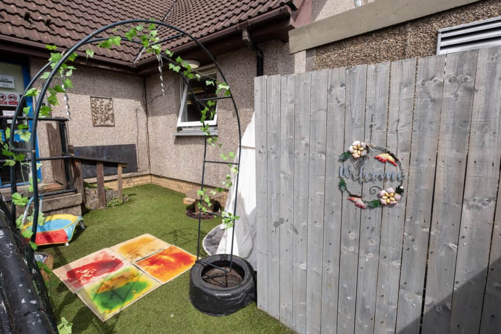 Bright outdoor play area at Thrive Childcare with artificial grass, colourful painting activity, welcome sign on wooden fence, and children's toys for early childhood development.
