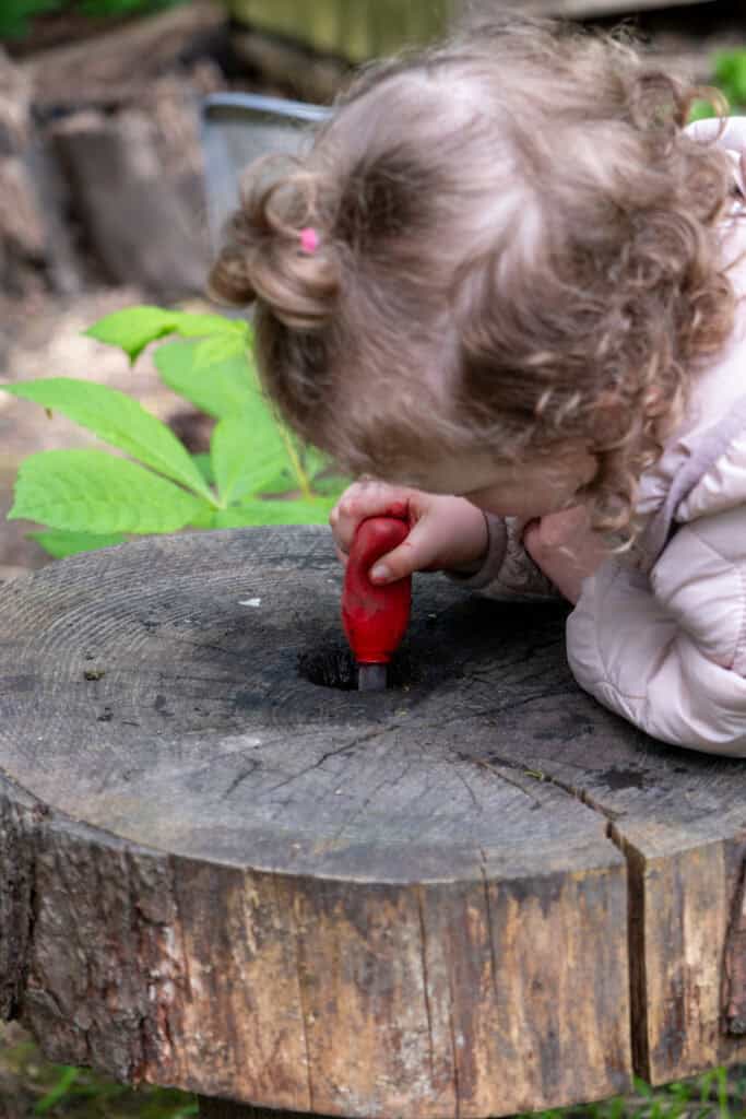 1. Child outdoors exploring nature with a red toy tool on a tree stump, part of Thrive Childcare’s outdoor learning environment for early childhood development.