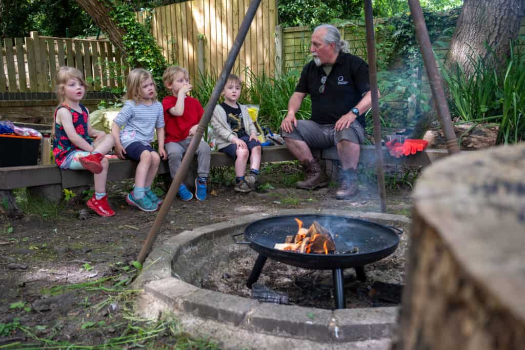 Woodland outdoor learning with children and teacher sitting by campfire at Thrive Childcare, promoting nature-based education and outdoor activities for early childhood development.