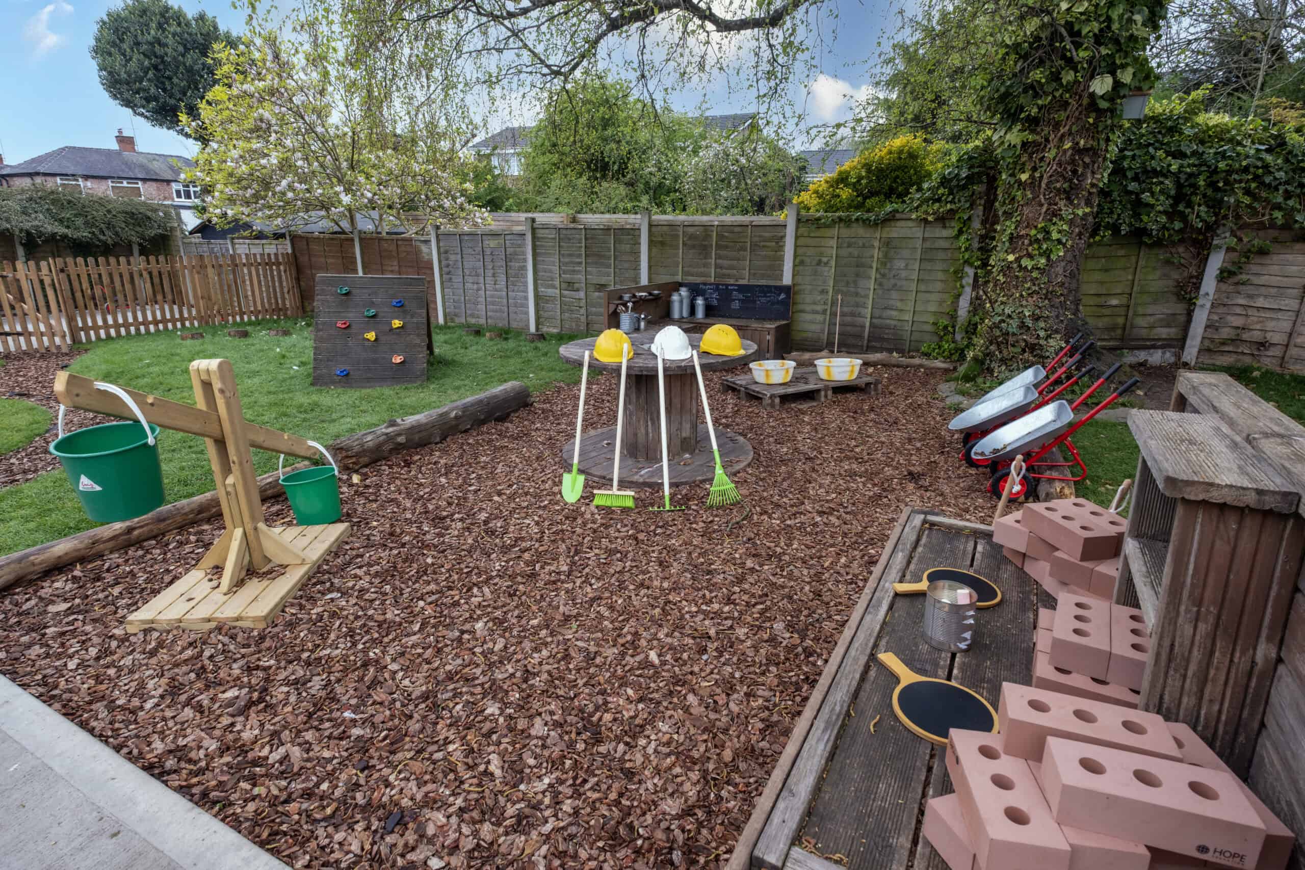 A colourful and outdoor play area for children at Thrive Childcare featuring a garden, playground equipment, climbing wall, seating, and gardening tools for early childhood development and outdoor fun.