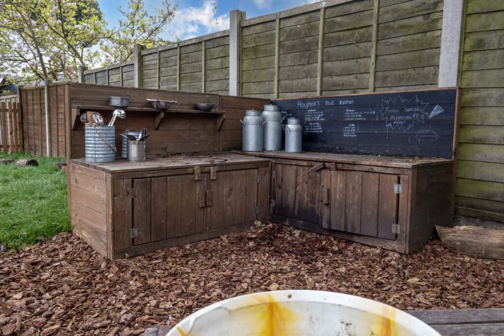 A children's outdoor mud kitchen with wooden counters, metal milk jugs, and chalkboard signs, perfect for early childhood play and learning at Thrive Childcare.