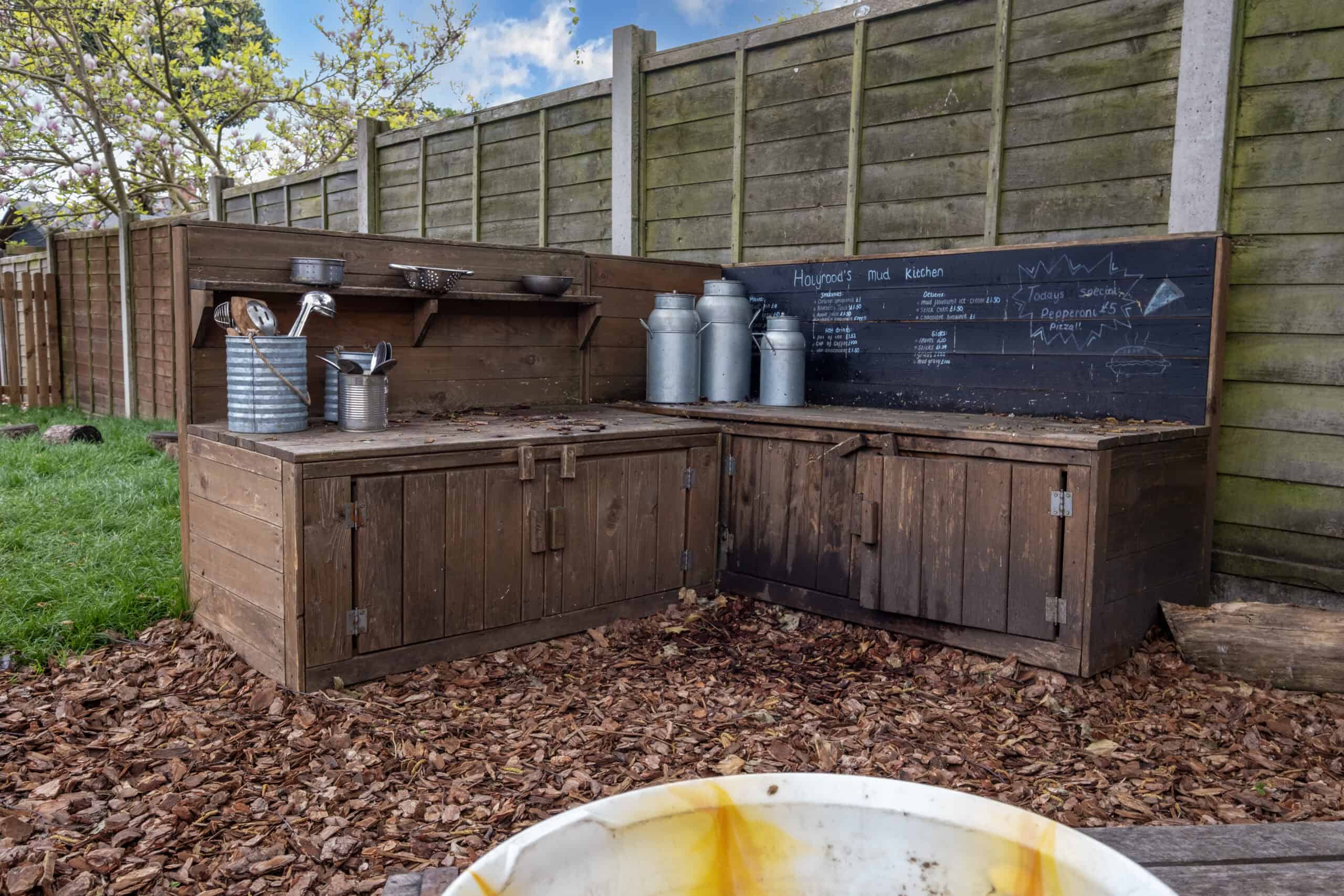 A children's outdoor mud kitchen with wooden counters, metal milk jugs, and chalkboard signs, perfect for early childhood play and learning at Thrive Childcare.