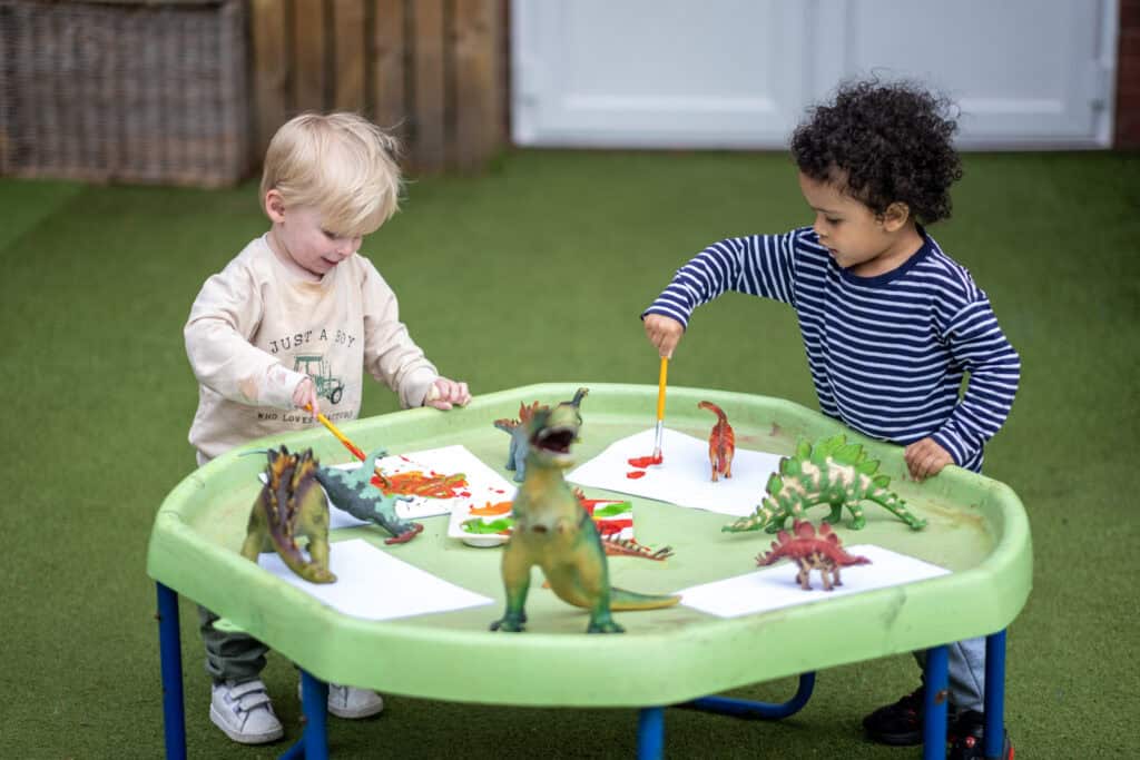 Bright young children playing creatively with dinosaur toys on a green outdoor activity table at Thrive Childcare, fostering early childhood development and imaginative play.