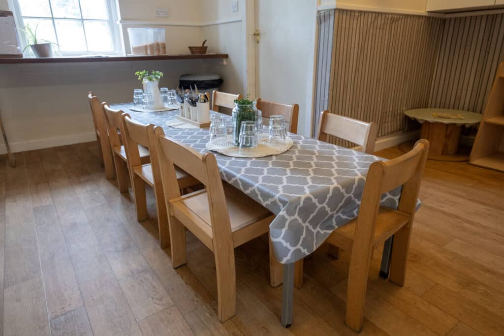 Soft natural light illuminating a warm, inviting children's daycare dining area with a wooden table set with glasses and greenery, promoting safe and engaging childcare environment.