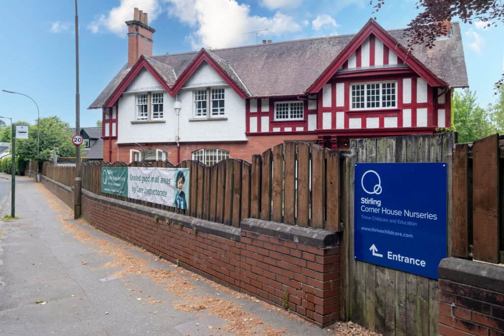 Bright red and white traditional Tudor-style nursery building, surrounded by a brick wall and wooden fencing, located in a residential area with street signage and clear blue sky.