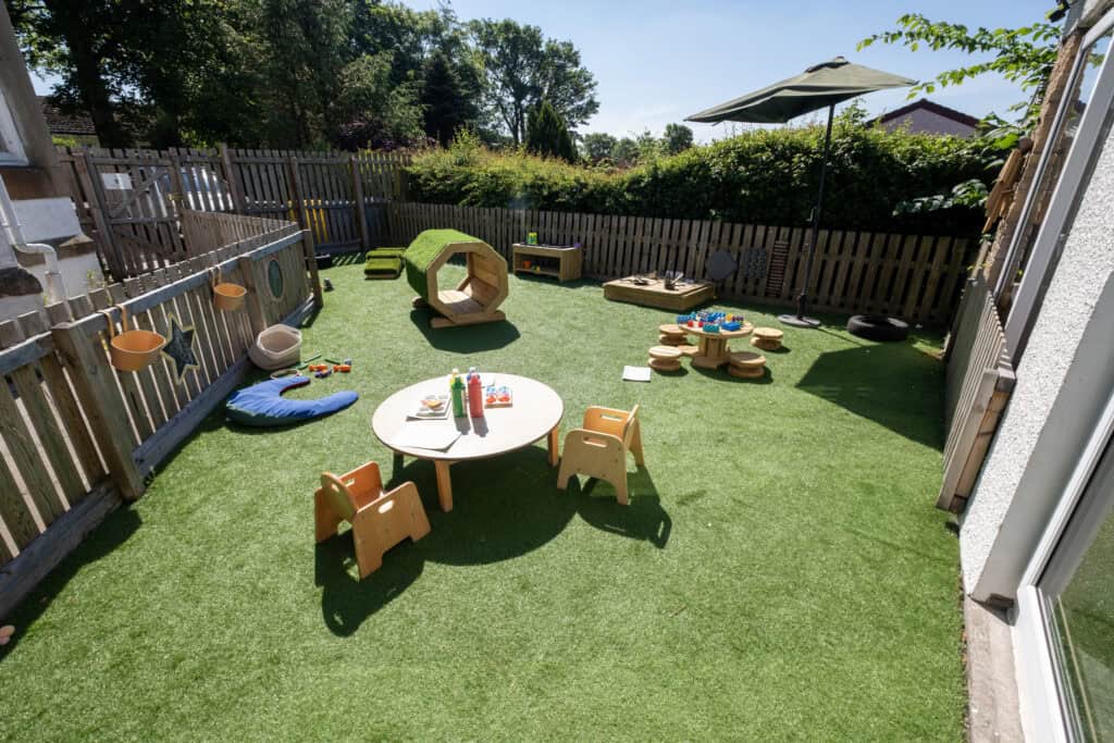 Bright outdoor childcare play area with artificial grass, toys, table, chairs, and shaded space, designed for early childhood development and outdoor learning at Thrive Childcare.