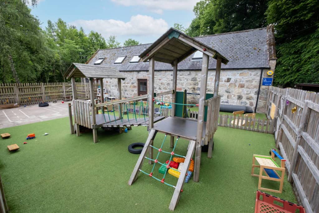 Bright outdoor playground at Thrive Childcare featuring wooden climbing structures, play equipment, soft turf, and a fenced area surrounded by lush green trees, ideal for early childhood play and development.