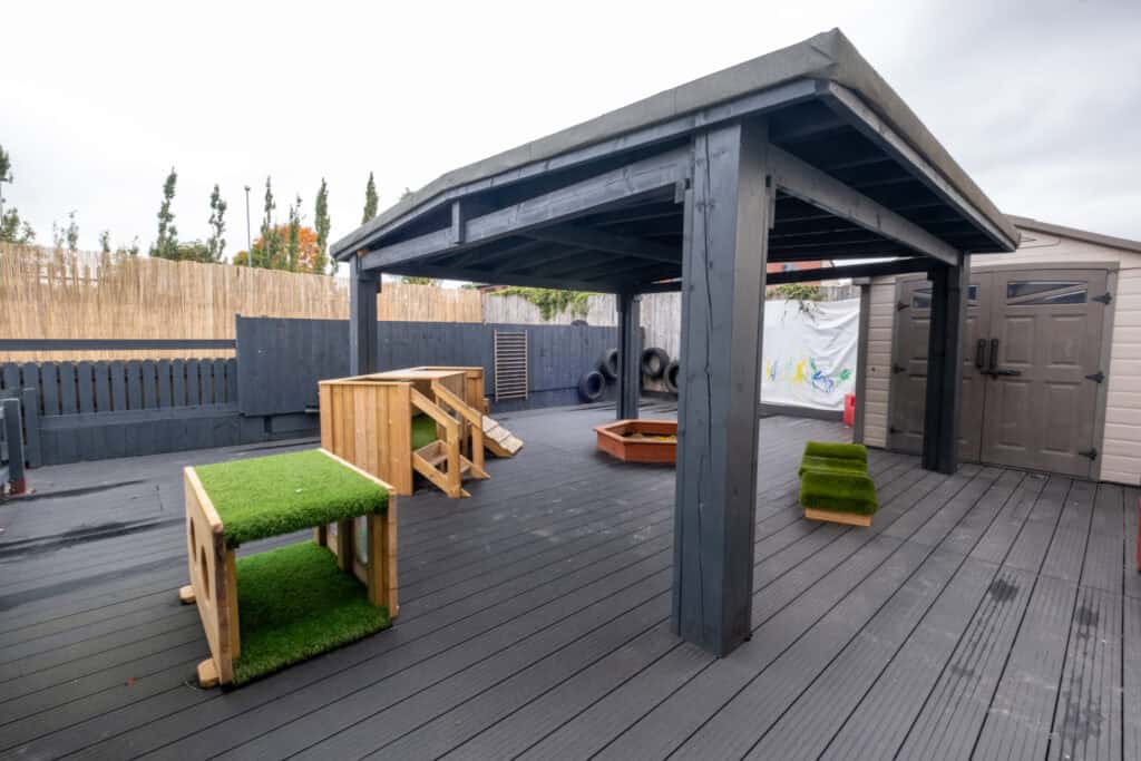 Bright outdoor playground with wooden play structures, artificial grass, and a shaded area at Thrive Childcare, offering a safe and engaging environment for young children to learn and play.