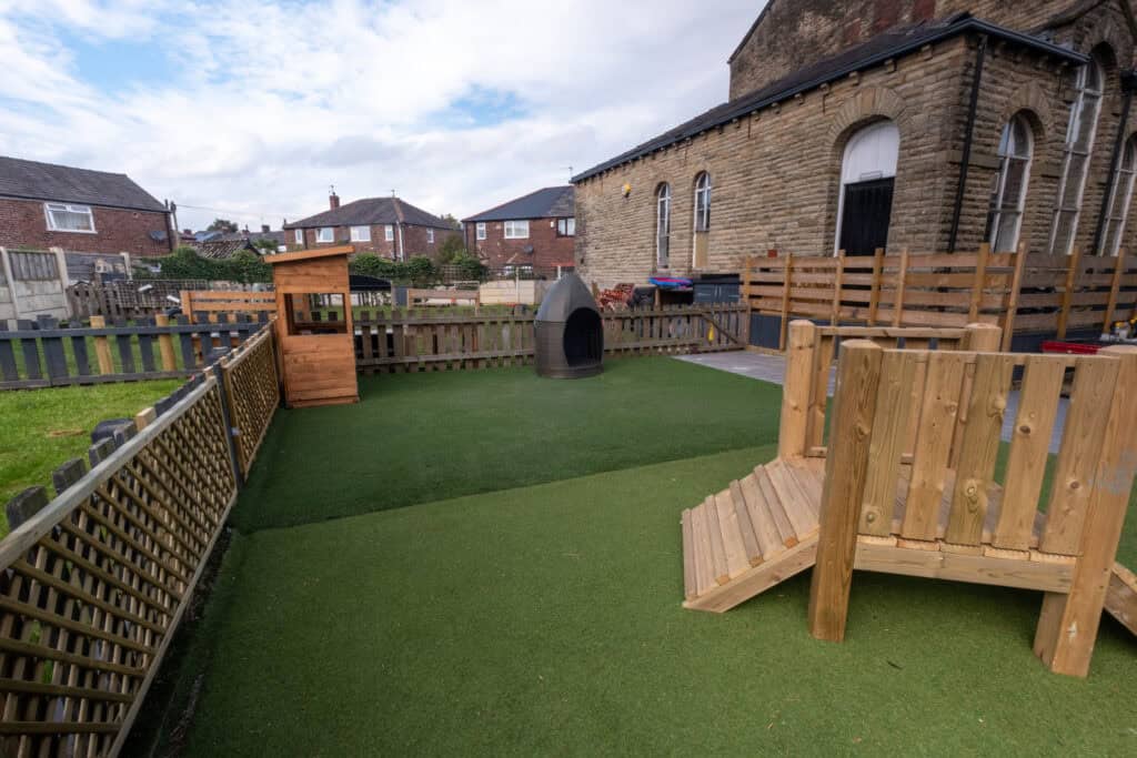 1. Bright outdoor childcare play area with artificial grass, wooden fencing, and child-friendly play equipment for early childhood development.