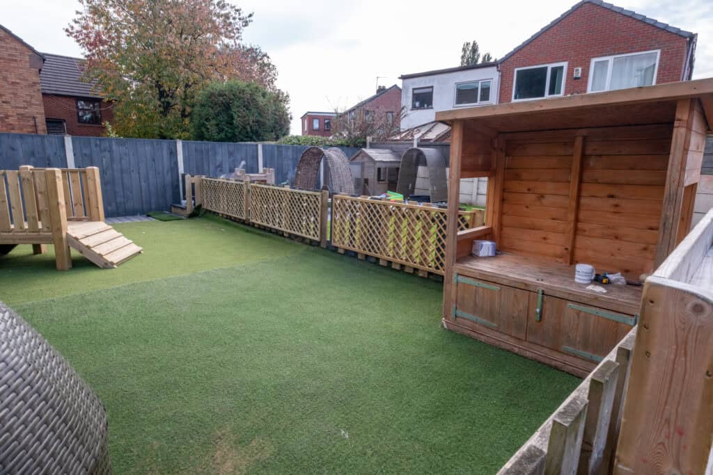 Bright outdoor childcare play area with artificial grass, wooden climbing and play structures, fenced for safety, ideal for early childhood development and outdoor learning at Thrive Childcare.