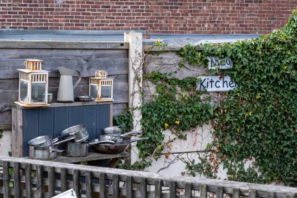 1. Outdoor kitchen area at Thrive Childcare with stainless steel pots, lanterns, and gardening tools, surrounded by greenery and a wooden fence for children’s outdoor activities.