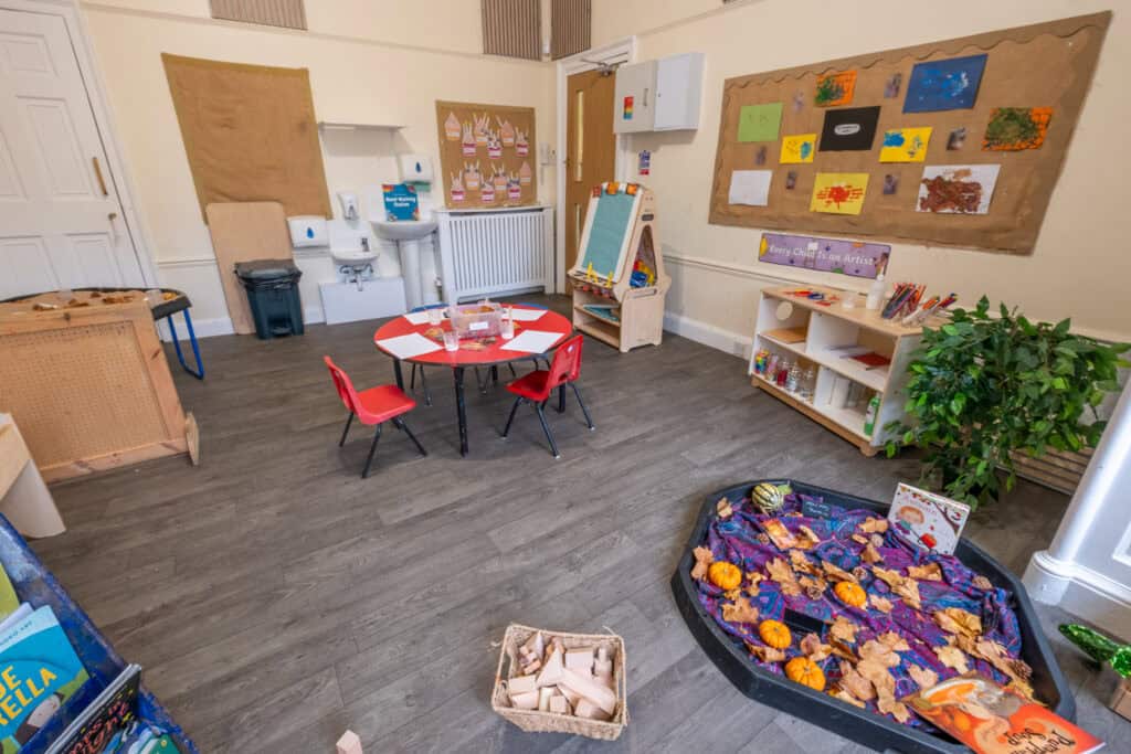 Bright and welcoming childcare room with colourful children's artwork, a round craft table with red chairs, educational toys, and reading materials, providing a nurturing environment for early childhood development.
