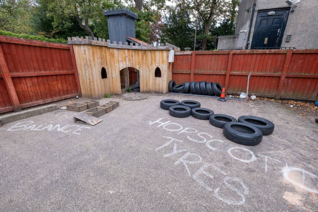 Colourful outdoor play area with tire obstacle course, chalk writings on pavement, wooden playhouse, and safety cones, designed for children's physical activity at Thrive Childcare.