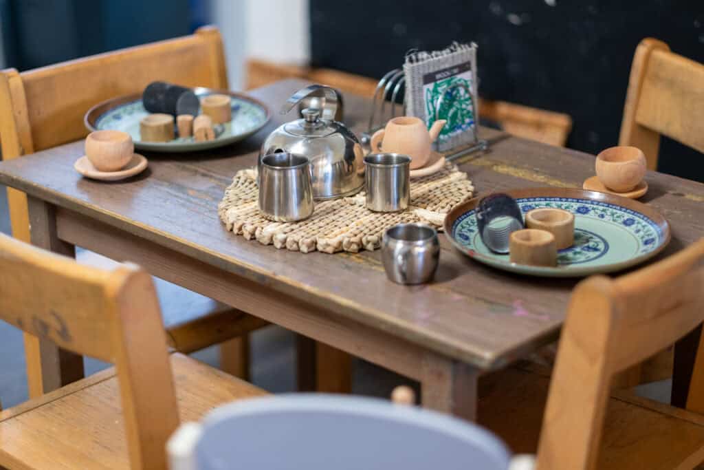 Delightful wooden child's dining table set with colourful ceramic plates, stainless steel cups, teapot, and wooden toy food, ideal for play-based learning and early childhood development at Thrive Childcare.