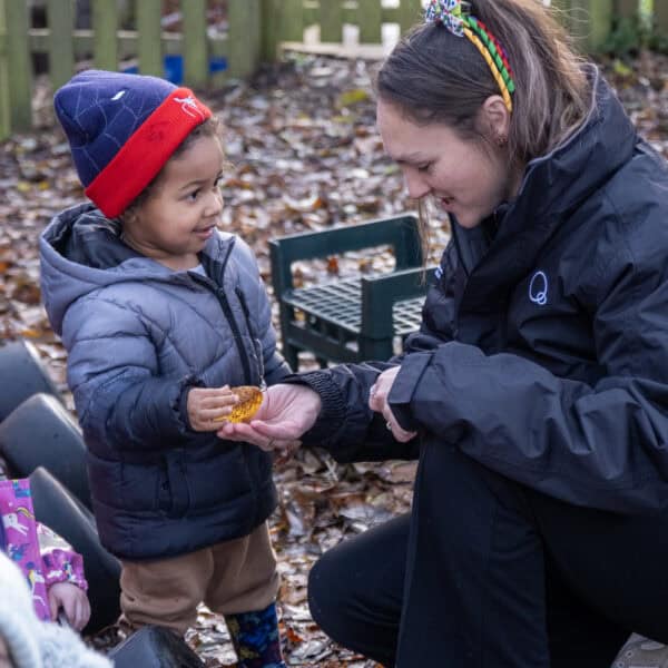 1. Child receiving a small gift from a caregiver outdoors at Thrive Childcare, in a woodland setting, promoting child development and outdoor learning.