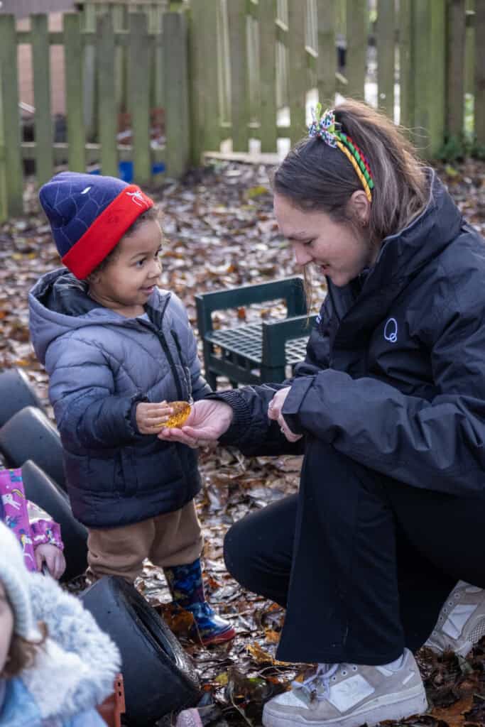 1. Child receiving a small gift from a caregiver outdoors at Thrive Childcare, in a woodland setting, promoting child development and outdoor learning.