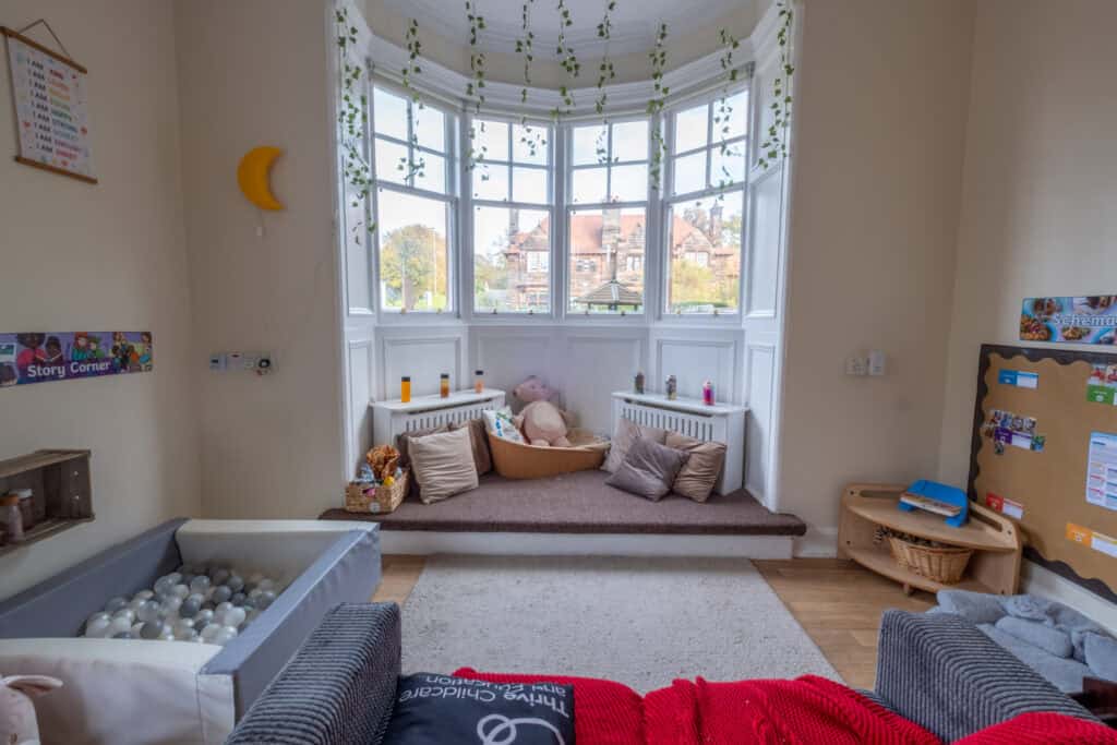 A bright, welcoming baby playroom with soft seating, toys, and a window nook with cushions, ideal for early childhood development and engaging activities at Thrive Childcare.