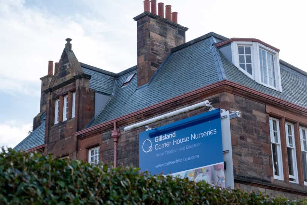 Bright blue sign for Gillsland Corner House Nurseries at Thrive Childcare, set against a historic stone building with slate roof and multiple chimneys, promoting childcare and early education services.