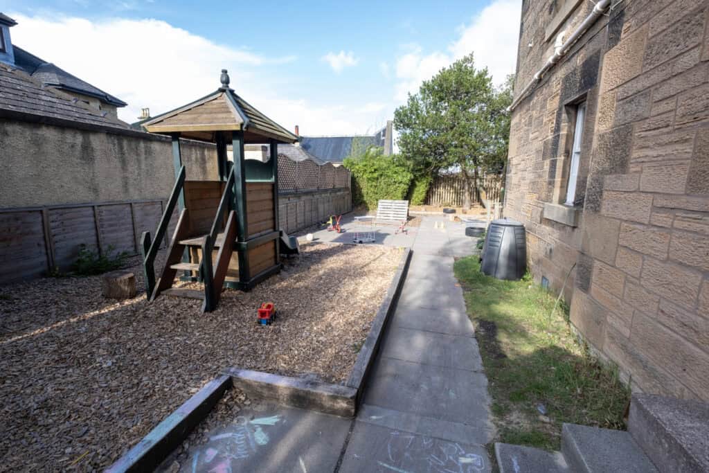 Colourful outdoor play area at Thrive Childcare with a wooden playhouse, toys, and shaded space ideal for children's early years development in a safe, welcoming environment.