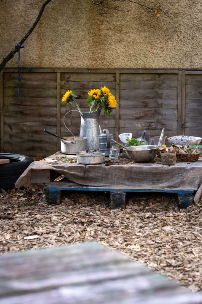 Bright sunflower arrangement in a watering can with children's outdoor play area, natural wooden fence, and wood chip ground, promoting outdoor learning and play for toddlers at Thrive Childcare.