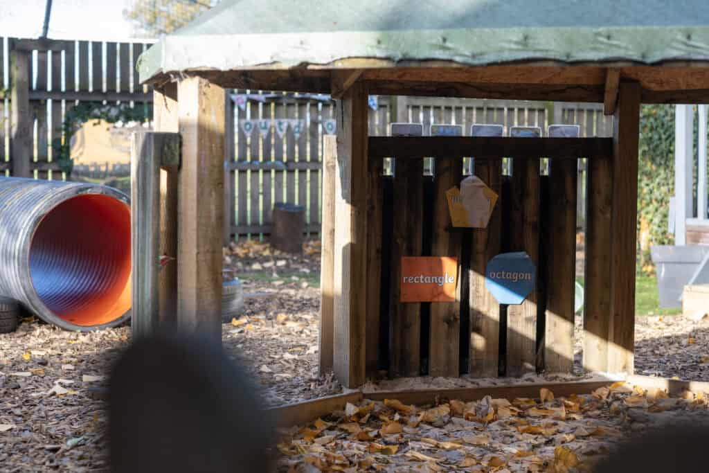 Colourful outdoor wooden play house at Thrive Childcare with educational shapes and autumn leaves on the ground, offering children a stimulating natural environment for outdoor learning and play.