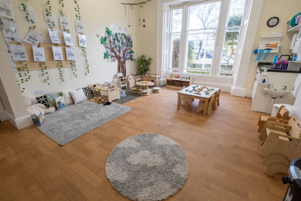 Bright, welcoming childcare classroom with wooden flooring, diverse play areas, educational displays, and natural light, perfect for early childhood development and learning activities.