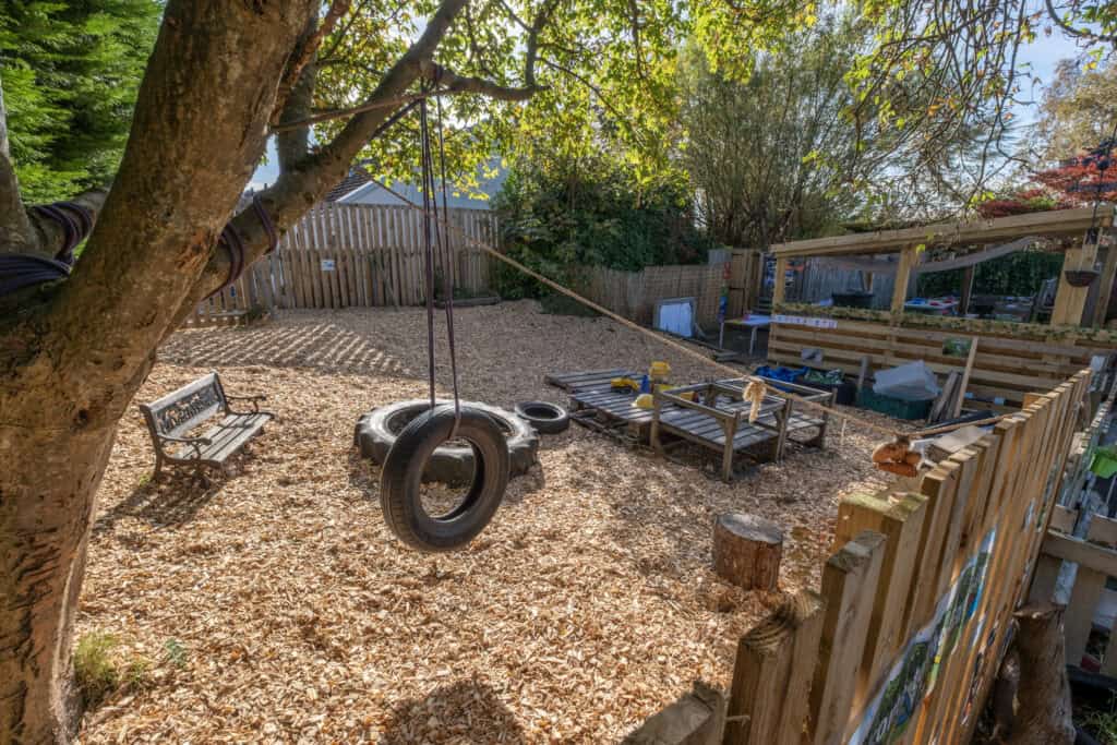 A lush outdoor children's play area at Thrive Childcare with a tire swing hanging from a tree, wooden benches, and fenced garden space for imaginative and active play.