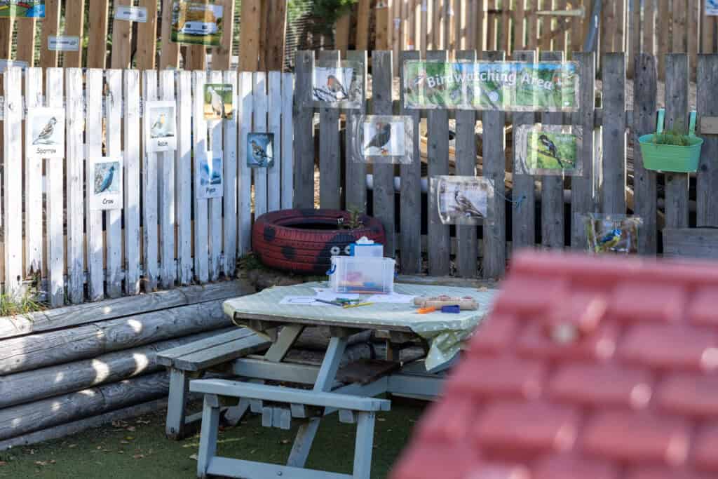 Bright outdoor birdwatching area at Thrive Childcare with educational bird photos, a picnic table, and natural wooden fencing, offering children engaging nature experiences and learning opportunities.