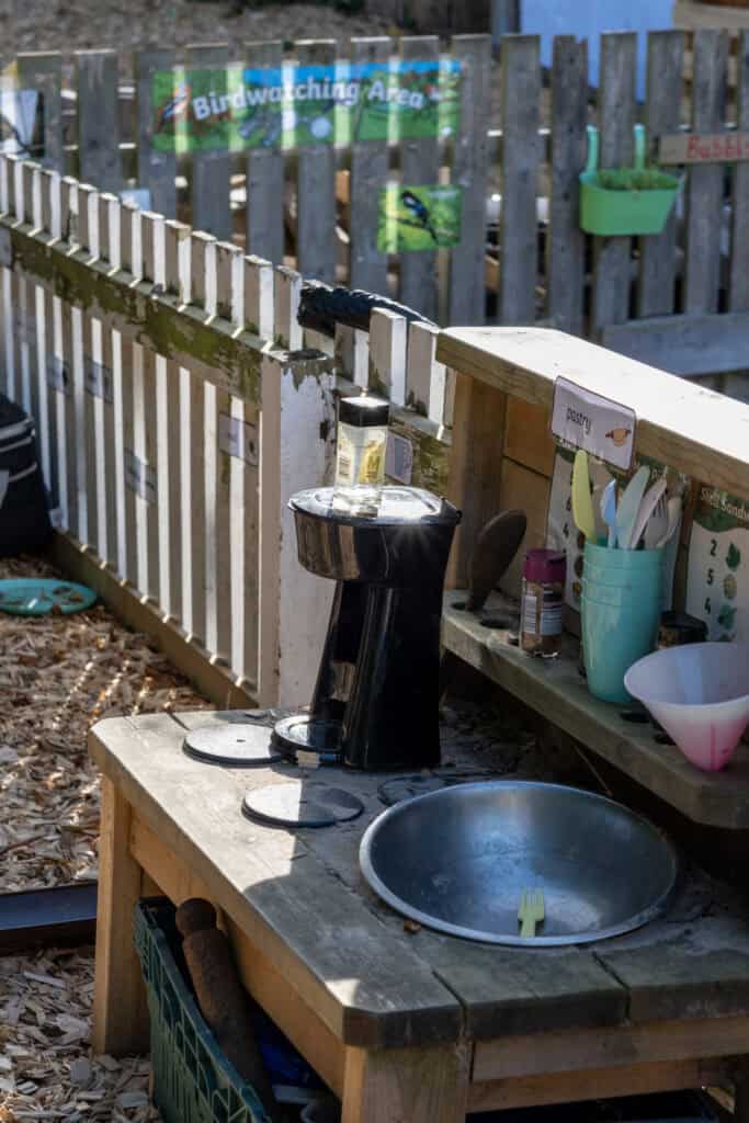 1. Outdoor children's play area with a birdwatching station, wooden fencing, and child-friendly kitchen play setup for preschool learning.