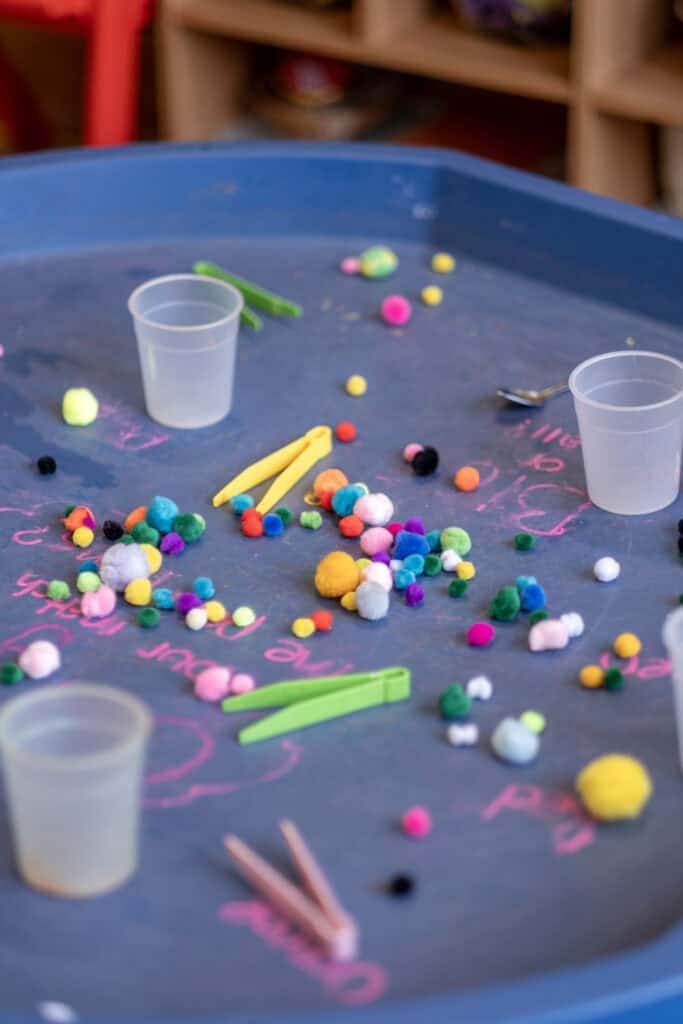 Bright, playful learning activity at Thrive Childcare featuring colourful pom-poms, chalk drawings, and child-friendly tools on a chalkboard tabletop to stimulate creativity and development in early childhood care.