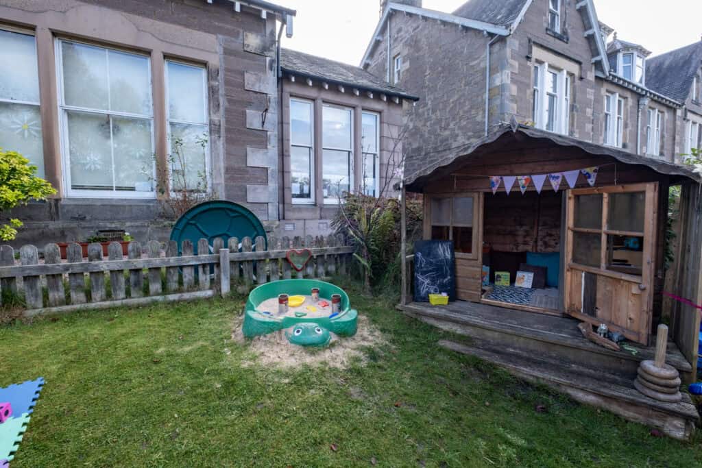 Bright outdoor play area at Thrive Childcare featuring a wooden playhouse, sandpit, and secure fencing for safe, engaging early years education and childcare in a friendly environment.