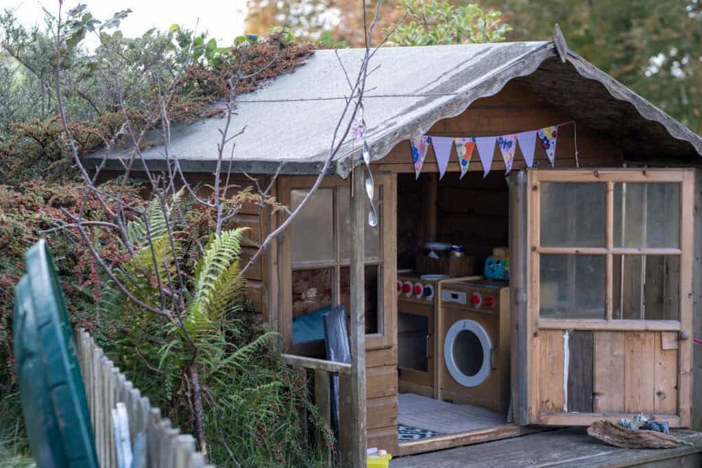 Bright wooden outdoor playhouse with children's toys, laundry, and colourful bunting, ideal for imaginative play in a childcare setting. Perfect for promoting outdoor learning and creativity.