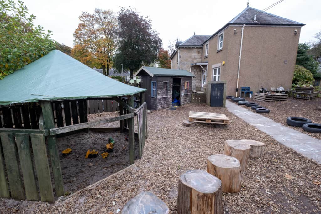 Bright outdoor play area at Thrive Childcare, featuring natural woodland surroundings, wooden play structures, tire stepping stones, and ample space for outdoor activities and exploration.