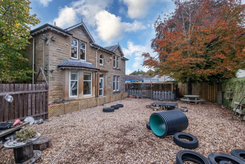 A large outdoor childcare play area with natural wood chips, tires, and playground equipment surrounded by a wooden fence, with a historic stone building and colourful autumn trees in the background.