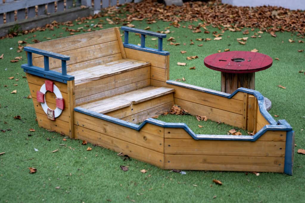 A colourful wooden boat-shaped play area with steps and a small round table, set on a lush green outdoor lawn with fallen autumn leaves at Thrive Childcare. Perfect for children's outdoor activities.