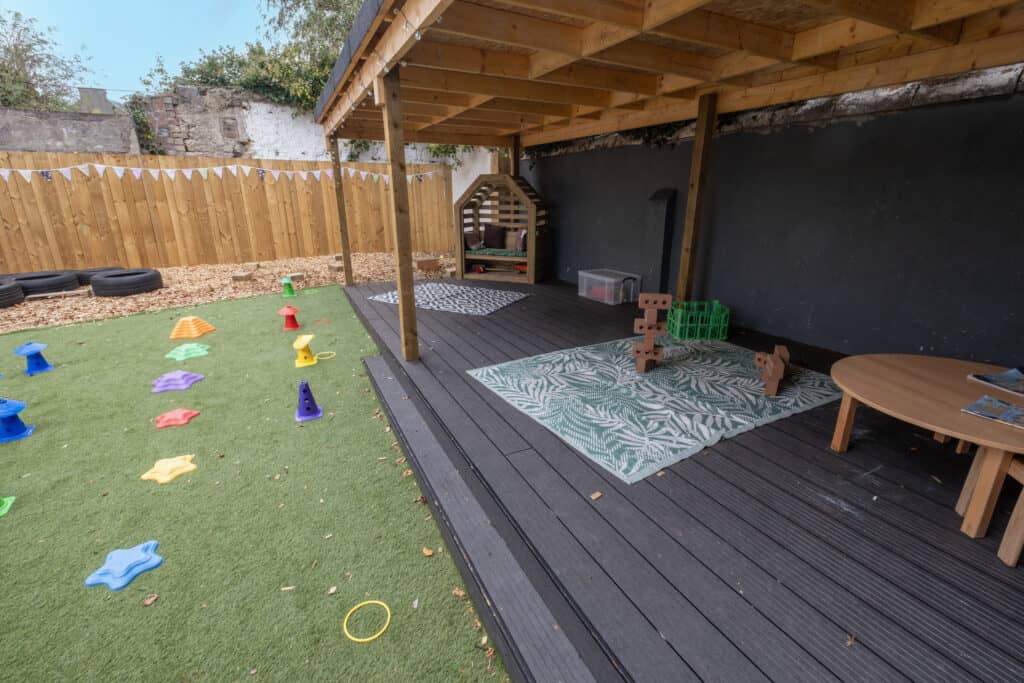 Bright outdoor toddler play area with colourful climbing toys, shaded seating, and artificial grass, offering a safe, engaging environment for early childhood development and outdoor learning.