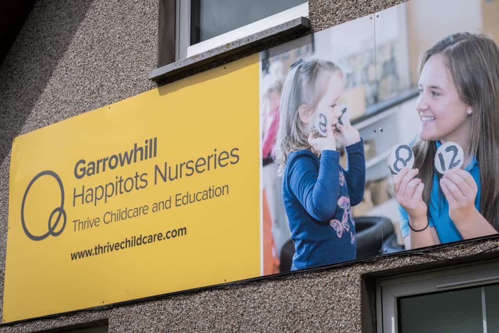 Bright yellow sign for Garrowhill Happitots Nurseries, promoting childcare and education services; featuring a happy young girl and caregiver engaging in educational activities.