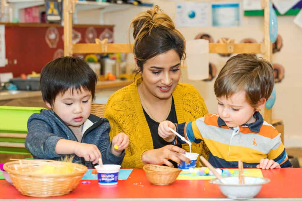 Brightly coloured classroom with a caring adult engaging young children in arts and crafts, fostering creativity and early childhood development.