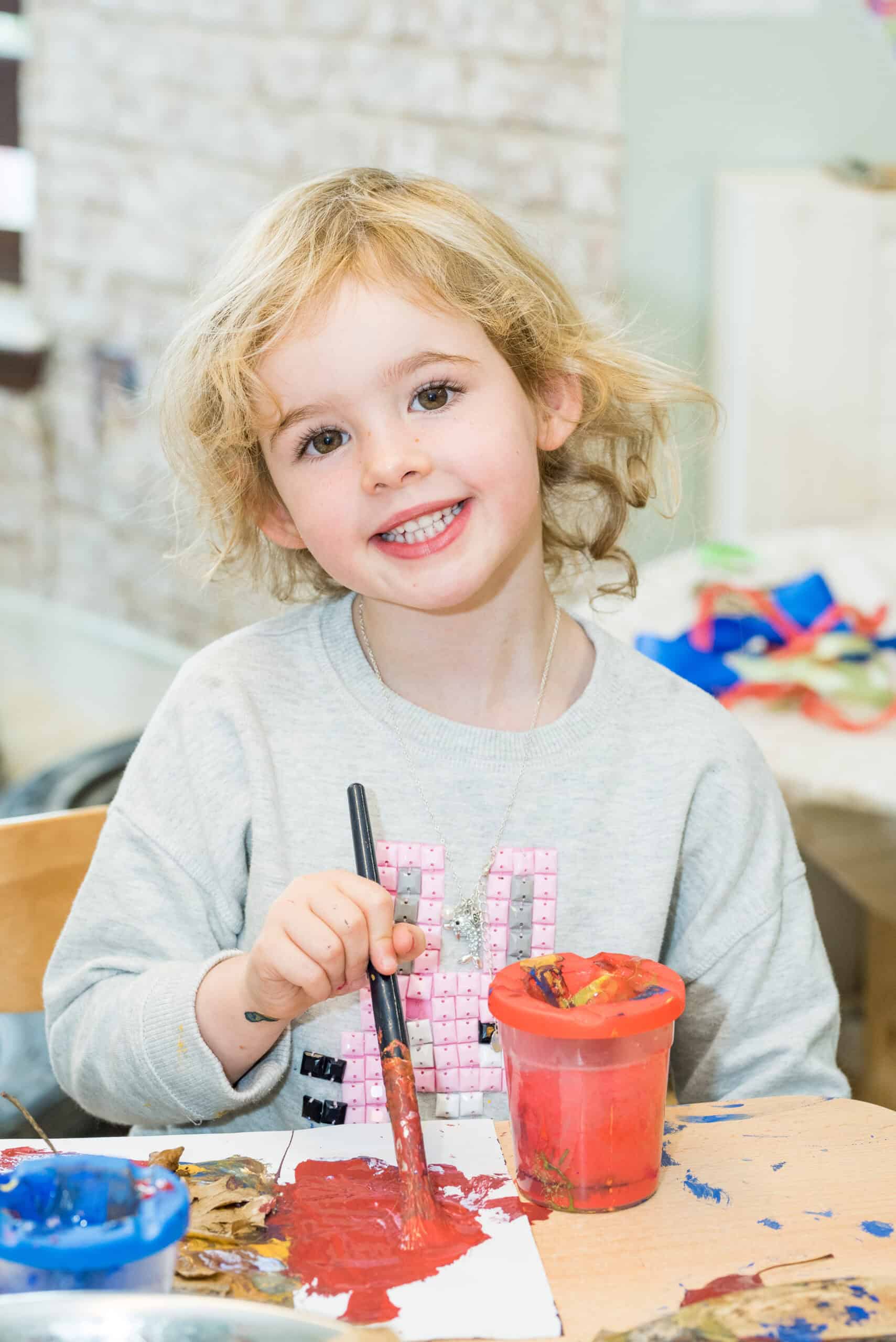 1. Happy young girl painting with red paint at preschool, showcasing creative early childhood education and caring childcare services in the UK.