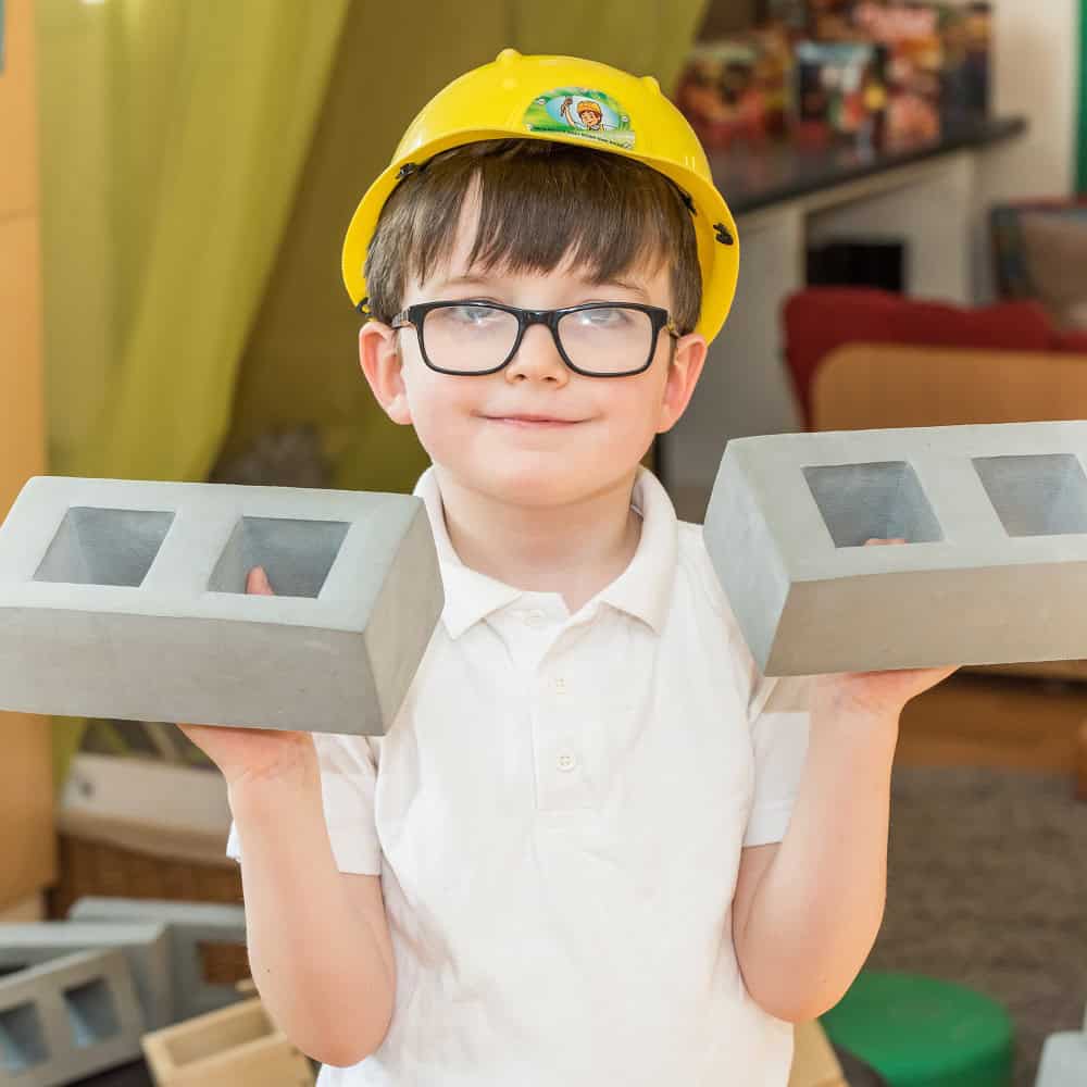 1. Young boy wearing a yellow safety helmet and glasses, holding toy concrete blocks, in a colourful childcare setting.